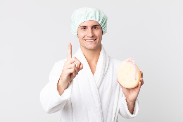 jovem bonito sorrindo com orgulho e confiança tornando-se o número um com roupão de banho, touca de banho e uma esponja