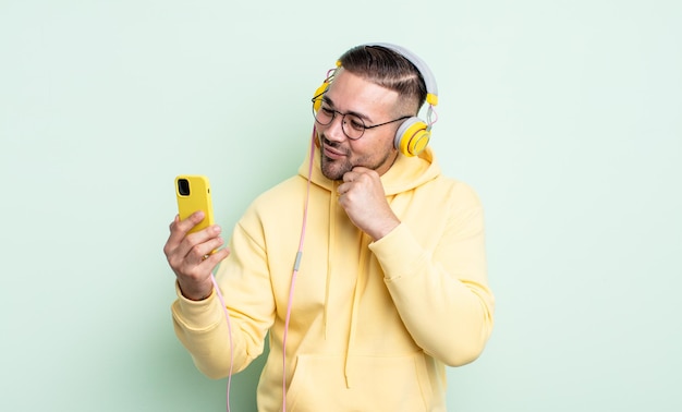 Jovem bonito sorrindo com uma expressão feliz e confiante com a mão no queixo. conceito de fones de ouvido e smartphone