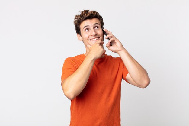 Jovem bonito sorrindo com uma expressão feliz e confiante, com a mão no queixo e falando com um telefone inteligente