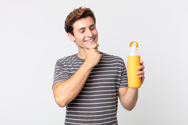 jovem bonito sorrindo com uma expressão feliz e confiante com a mão no queixo e segurando uma garrafa térmica de café