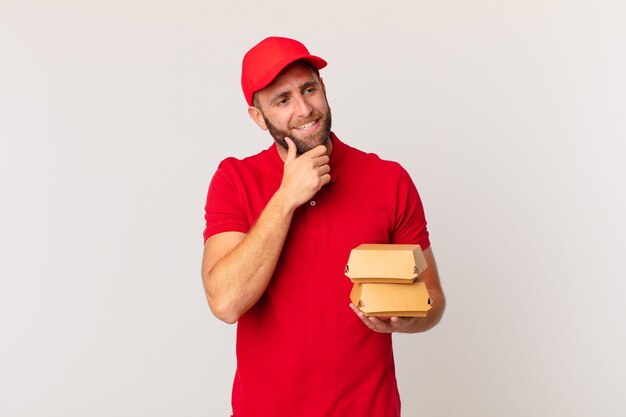 Jovem bonito sorrindo com uma expressão feliz e confiante com a mão no queixo entregando um hambúrguer conceito