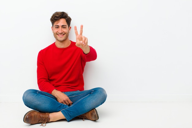 Jovem bonito sorrindo e olhando feliz, despreocupado e positivo, gesticulando vitória ou paz com uma mão sentada no chão