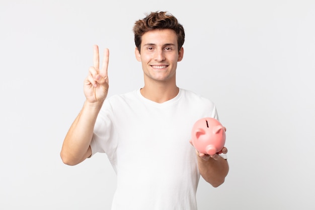 Jovem bonito sorrindo e parecendo amigável, mostrando o número dois e segurando um cofrinho