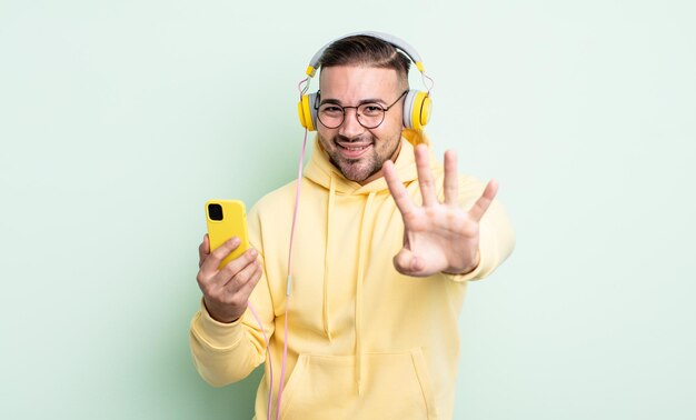 jovem bonito sorrindo e parecendo amigável, mostrando o número quatro. fones de ouvido e conceito de smartphone