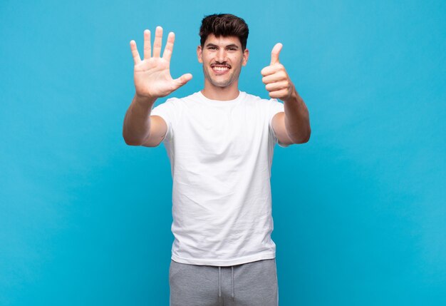 Jovem bonito sorrindo e parecendo amigável, mostrando o número seis ou sexto com a mão para a frente, em contagem regressiva