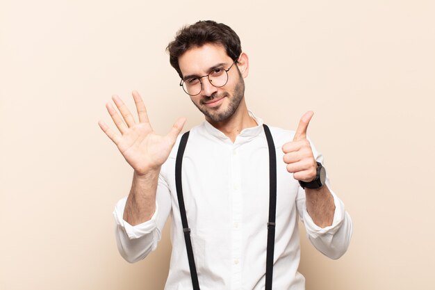 Jovem bonito sorrindo e parecendo amigável, mostrando o número seis ou sexto com a mão para a frente, em contagem regressiva