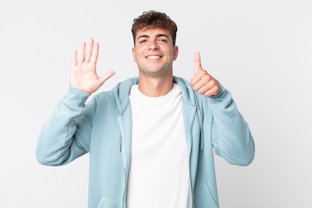 Jovem bonito sorrindo e parecendo amigável, mostrando o número seis ou sexto com a mão para a frente, em contagem regressiva