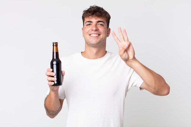 Jovem bonito sorrindo e parecendo amigável, mostrando o número três e segurando uma garrafa de cerveja