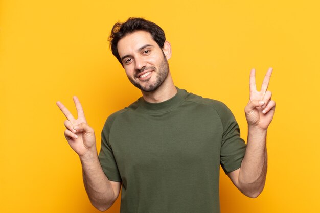 Jovem bonito sorrindo e parecendo feliz, amigável e satisfeito, gesticulando vitória ou paz com as duas mãos