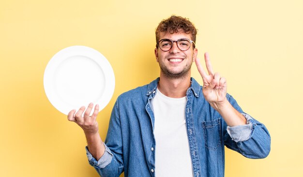 Jovem bonito sorrindo e parecendo feliz, gesticulando vitória ou paz. conceito de prato vazio