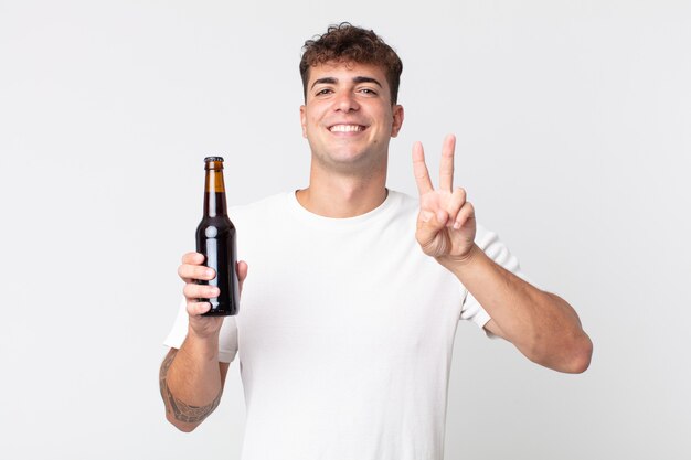 Jovem bonito sorrindo e parecendo feliz, gesticulando vitória ou paz e segurando uma garrafa de cerveja