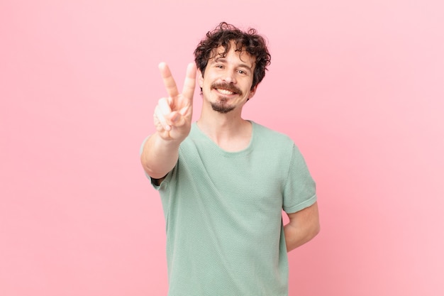 Jovem bonito sorrindo e parecendo feliz, gesticulando vitória ou paz