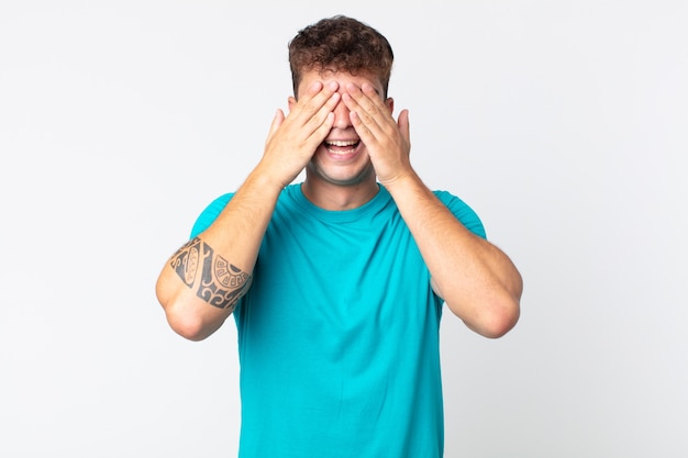 Jovem bonito sorrindo e se sentindo feliz, cobrindo os olhos com as duas mãos e esperando por uma surpresa incrível