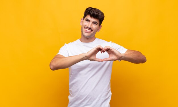 Jovem bonito sorrindo e se sentindo feliz, fofo, romântico e apaixonado, fazendo formato de coração com as duas mãos