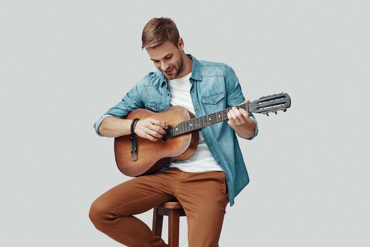 Foto jovem bonito sorrindo e tocando guitarra enquanto está sentado contra um fundo cinza