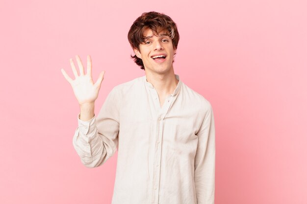 Jovem bonito sorrindo feliz, acenando com a mão, dando as boas-vindas e cumprimentando você