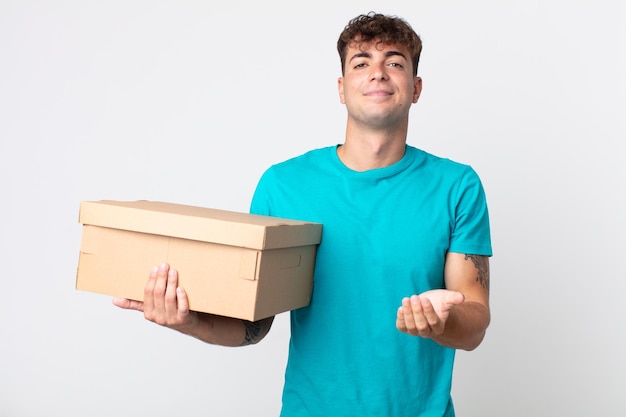 Jovem bonito sorrindo feliz com simpatia e oferecendo e mostrando um conceito e segurando uma caixa de papelão