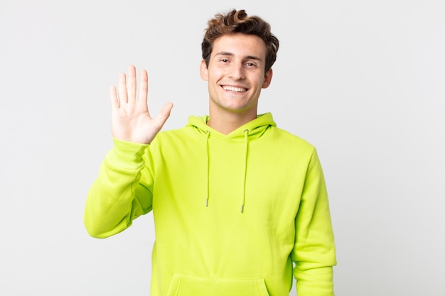 Jovem bonito sorrindo feliz e alegre, acenando com a mão, dando as boas-vindas e cumprimentando você ou dizendo adeus