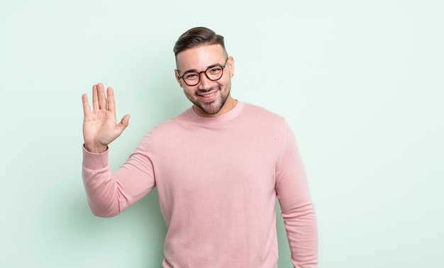 Jovem bonito sorrindo feliz e alegre, acenando com a mão, dando as boas-vindas e cumprimentando você ou dizendo adeus