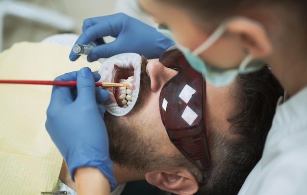 Jovem bonito sorrindo olhando a câmera dentista feminina verificando os dentes do paciente com espelho na clínica odontológica moderna examinar o jovem pelo dentista na luz de fundo desfocada