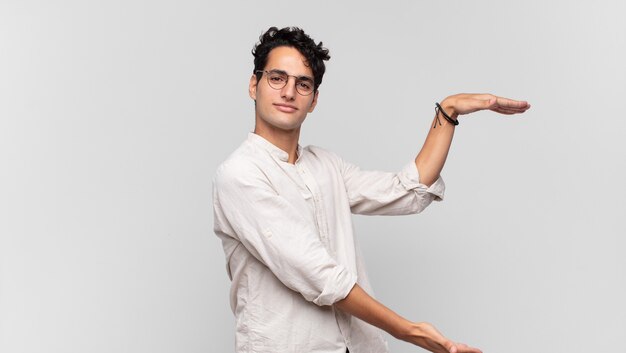 Jovem bonito sorrindo, sentindo-se feliz, positivo e satisfeito, segurando ou mostrando o objeto ou conceito no espaço da cópia