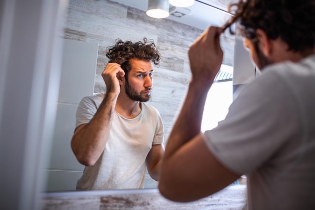 Foto jovem bonito tocando seu cabelo com a mão e aparando no banheiro em casa. homem branco metrossexual preocupado com a perda de cabelo e olhando no espelho sua linha do cabelo retrocedendo.