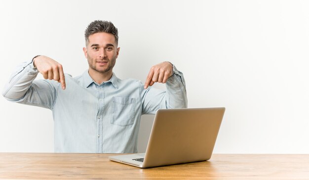 Jovem bonito trabalhando com seu laptop aponta para baixo com os dedos, sentimento positivo.