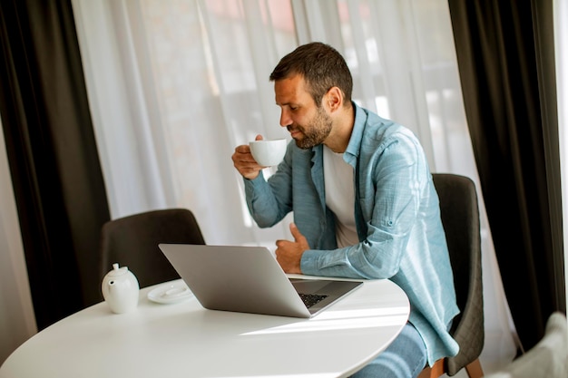 Jovem bonito usando laptop e bebendo chá na sala de estar