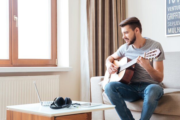 Foto jovem bonito usando laptop e tocando guitarra em casa