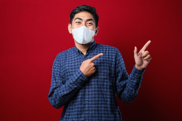 Jovem bonito usando máscara médica sorrindo e olhando para a câmera apontando com uma das mãos e dedos para o lado sobre fundo vermelho