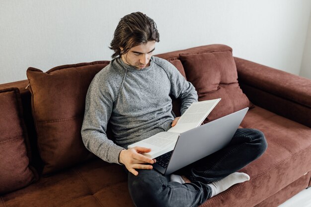 Jovem bonito usando o laptop em casa. Homem estudante está lendo o livro. Compras online, trabalho em casa, freelance, aprendizagem online, estudando o conceito. Educação a Distância