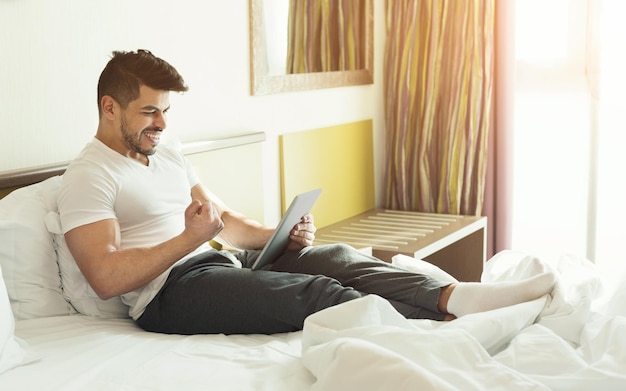 Jovem bonito usando tablet digital e celebrando o sucesso enquanto está sentado em uma cama em um quarto de hotel