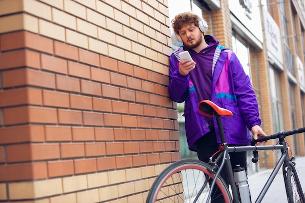 Jovem bonito usando telefone celular e fones de ouvido em pé perto de sua bicicleta ao lado dele
