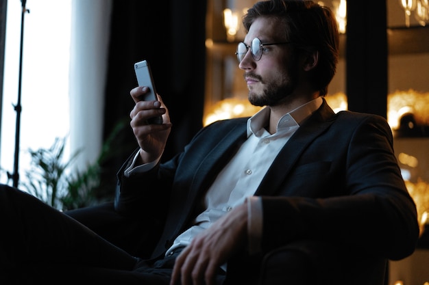 Jovem bonito usando telefone inteligente durante a noite. Closeup rosto de empresário feliz mensagens no celular à noite no café.