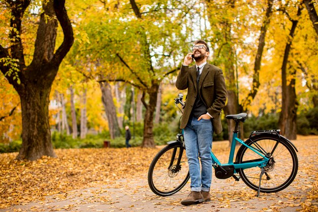 Jovem bonito usando um telefone celular em uma bicicleta elétrica no parque outono