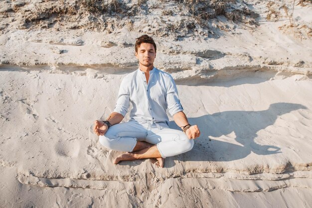 Jovem bonito vestido com roupas leves sentado em pose de meditação na praia de areia