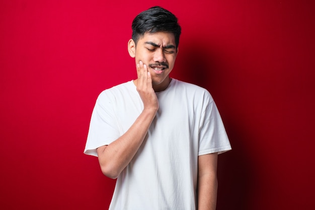 Jovem bonito vestindo camiseta branca em pé sobre fundo vermelho isolado, tocando a boca com a mão com expressão dolorosa por causa de dor de dente ou doença dentária nos dentes. Dentista