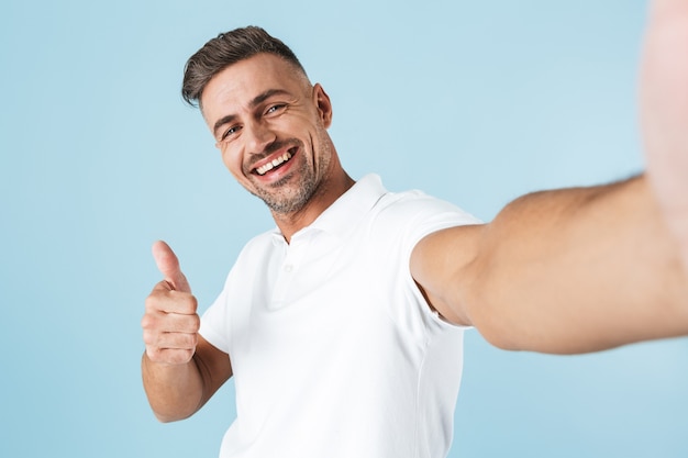 Jovem bonito vestindo camiseta branca em pé sobre o azul, tirando uma selfie e dando sinal de positivo