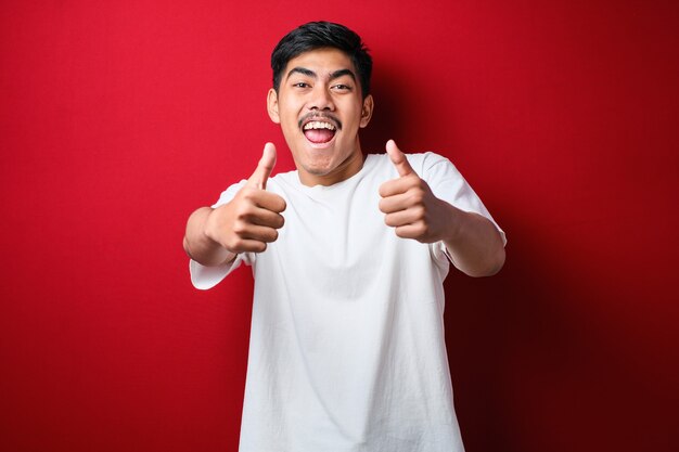 Jovem bonito vestindo camiseta branca sobre fundo vermelho, aprovando o gesto positivo com a mão, polegares para cima sorrindo e feliz pelo sucesso. Gesto do vencedor.