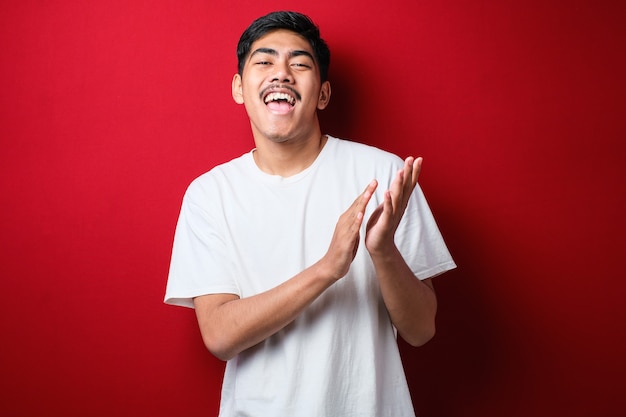 Jovem bonito vestindo camiseta casual sobre fundo vermelho batendo palmas e aplaudindo feliz e alegre, sorrindo orgulhosas mãos juntas