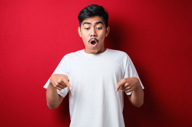 Jovem bonito vestindo uma camisa casual em pé sobre um fundo vermelho apontando para baixo com os dedos mostrando um anúncio, rosto surpreso e boca aberta