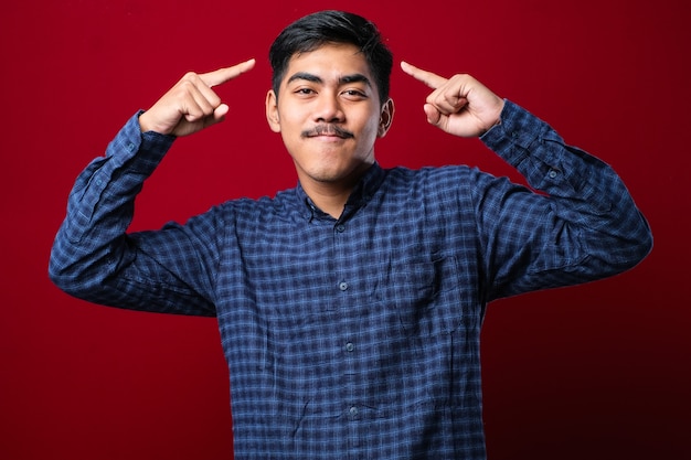 Jovem bonito vestindo uma camisa casual sorrindo apontando para a cabeça com os dedos das duas mãos, ótima ideia ou pensamento, boa memória sobre fundo vermelho