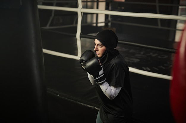 Jovem boxeadora séria em roupas ativas e hijab olhando para o saco de boxe