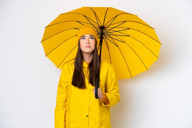 Jovem brasileira com casaco à prova de chuva e guarda-chuva isolado no fundo branco e olhando para cima