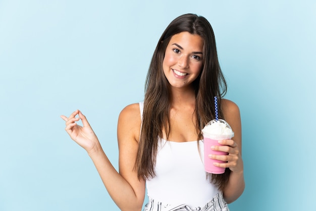 Jovem brasileira com milk-shake de morango isolado em um fundo azul apontando o dedo para o lado