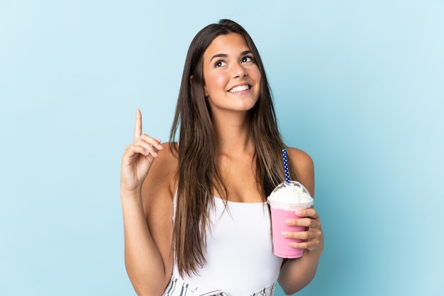 Jovem brasileira com milk-shake de morango isolado na parede azul apontando uma ótima ideia