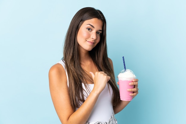 Jovem brasileira com milkshake de morango isolada em uma parede azul orgulhosa e satisfeita