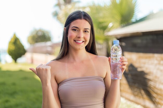 Jovem brasileira com uma garrafa de água ao ar livre apontando para o lado para apresentar um produto