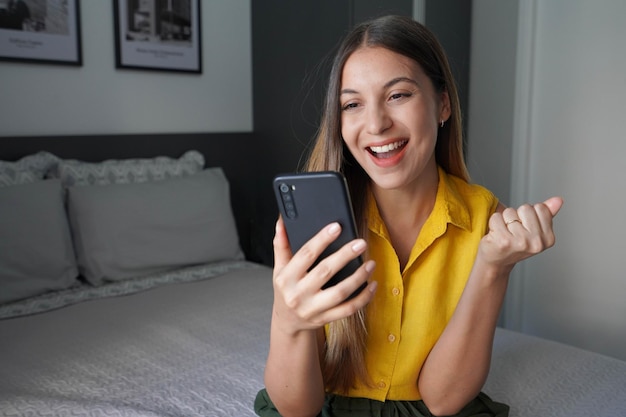 Jovem brasileira feliz segurando um smartphone e comemorando com o punho cerrado sentada na cama em casa