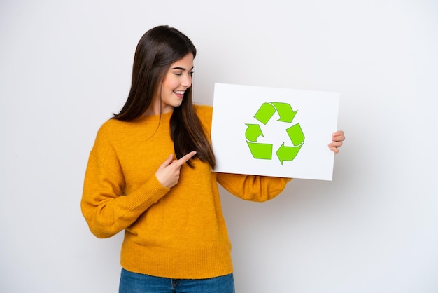 Jovem brasileira isolada em fundo branco segurando um cartaz com ícone de reciclagem e apontando-o